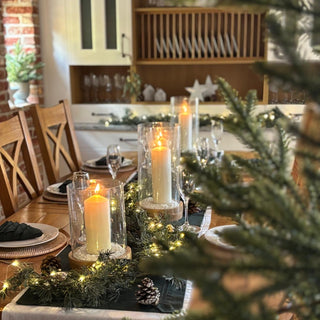 Dining table set for a festive meal with candles and greenery, kitchen in the background
