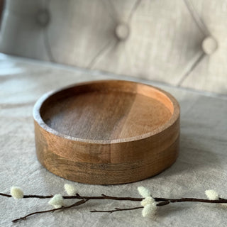 A round wooden candle plate  with a smooth, polished finish, displayed on a neutral fabric surface with a tufted chair in the background.