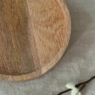 A round wooden candle plate with a natural wood grain finish, photographed from a top-down angle on a textured beige fabric surface with a small dried flower prop in the corner.