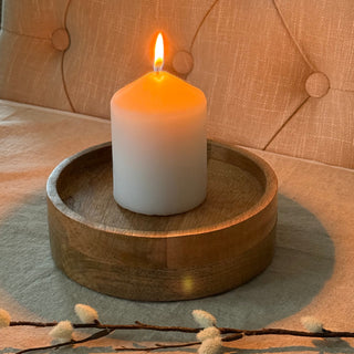 A lit white pillar candle sits in a round wooden candle plate on a beige upholstered chair, with soft decorative branches in the foreground.
